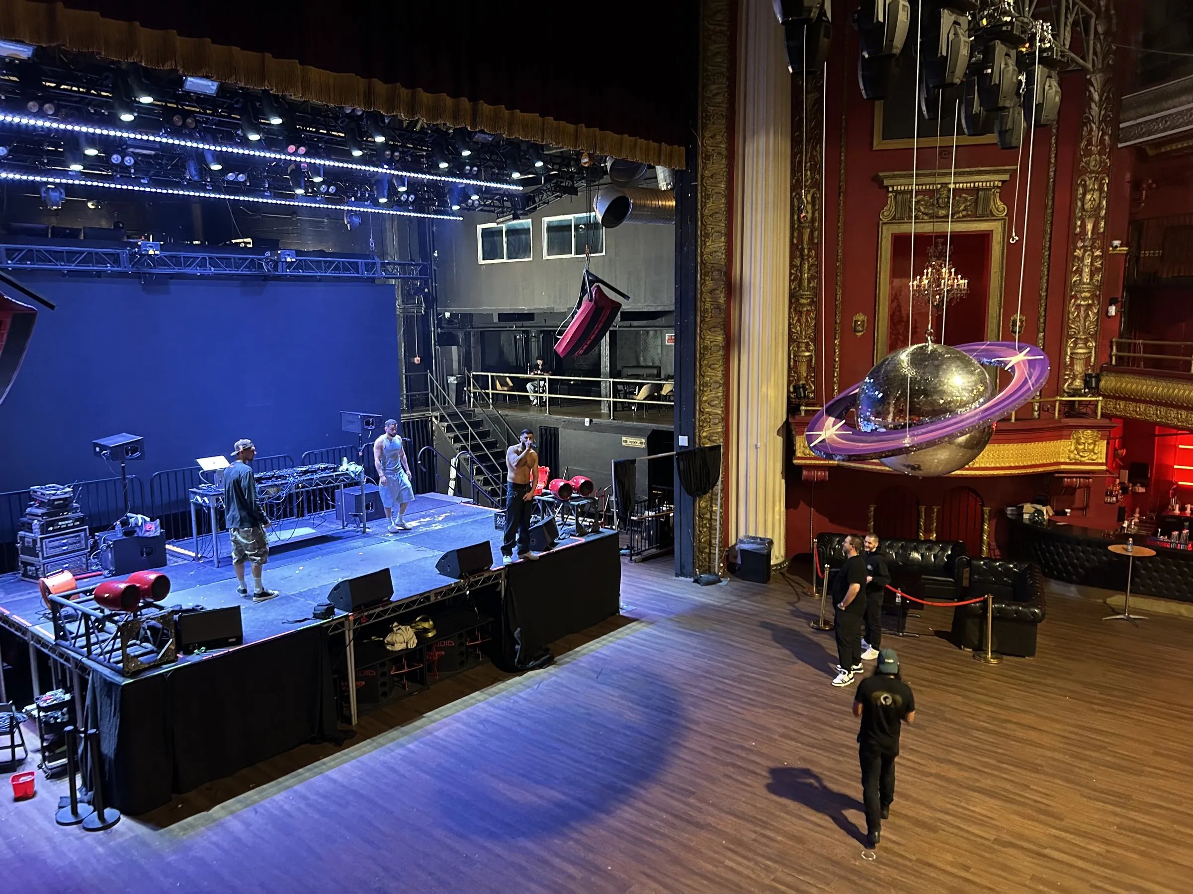 The theatre from the balcony, the stage lit blue with band gear set on the deck and a mirror ball ring hung over the floor.