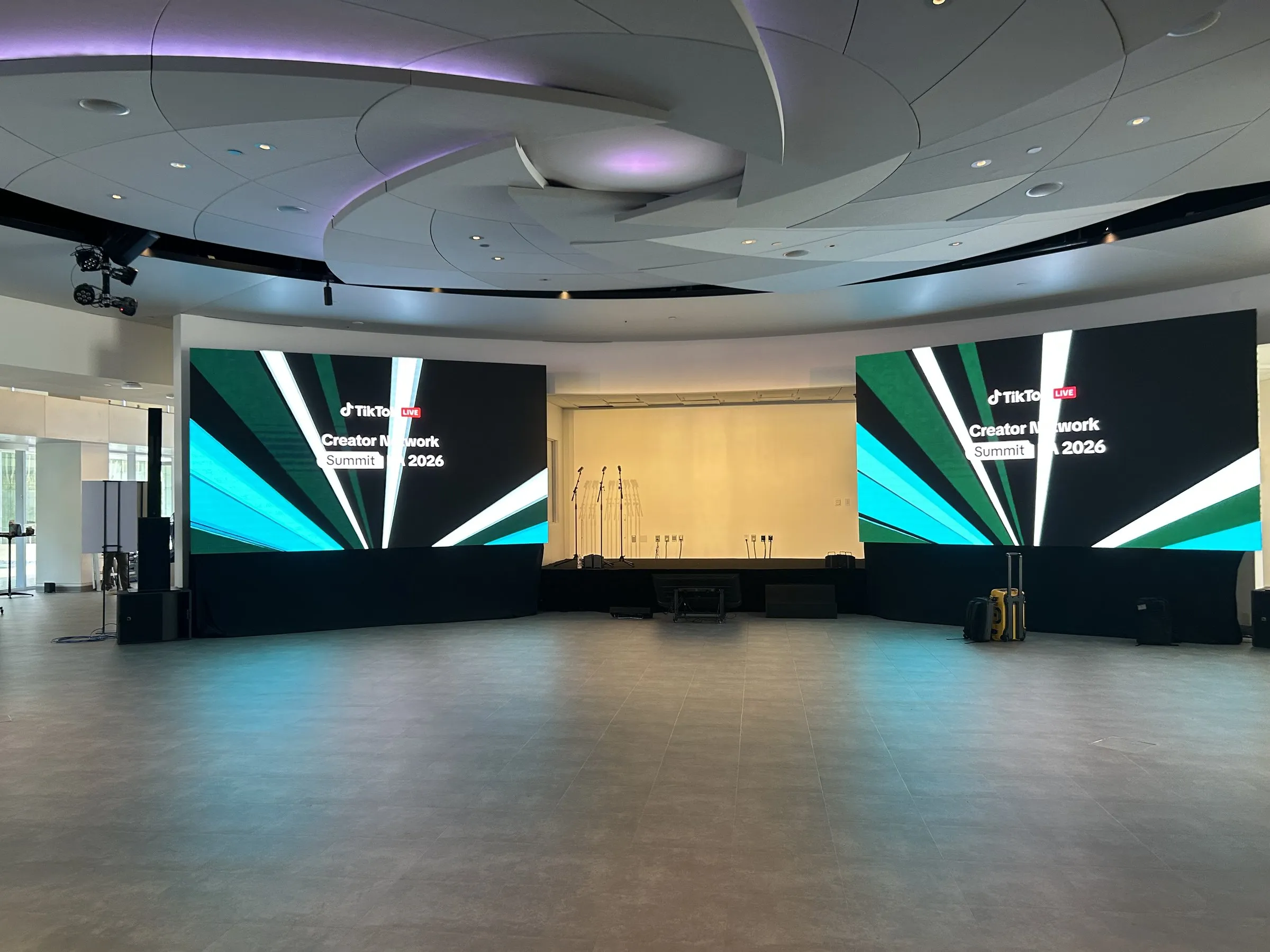 An empty pavilion room with a sculpted white ceiling, two LED walls flanking a projection screen, and a bare floor in front of the stage.